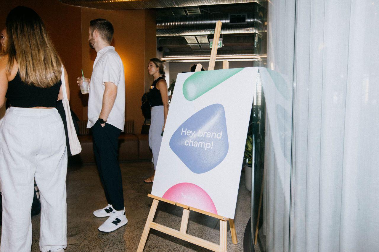 A group of marketing professionals gathered around an easel with a sign saying 'Hey brand champ!', discussing brand marketing strategies at a Tracksuit event