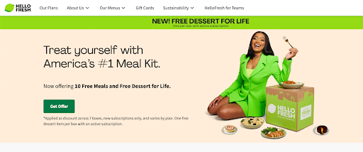 A professional woman in a green suit sitting at a table with a HelloFresh meal kit, representing the convenience and quality of HelloFresh's home-delivered meal service.