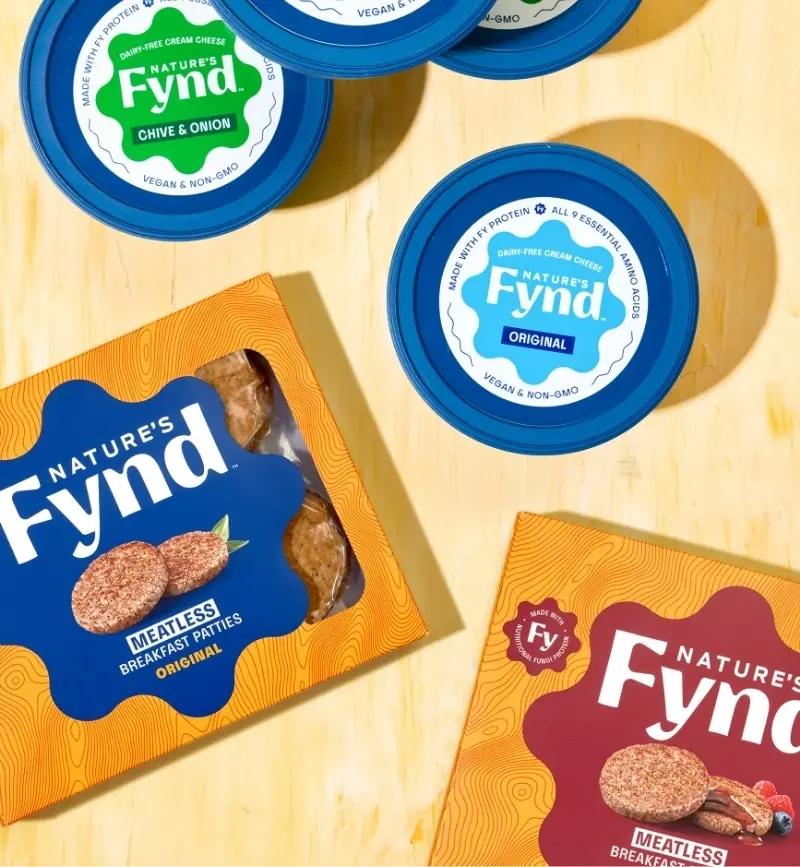 A variety of plant-based food products from Nature's Fynd displayed on a wooden table, representing the company's contribution to the sustainable and eco-friendly consumer packaged goods industry.