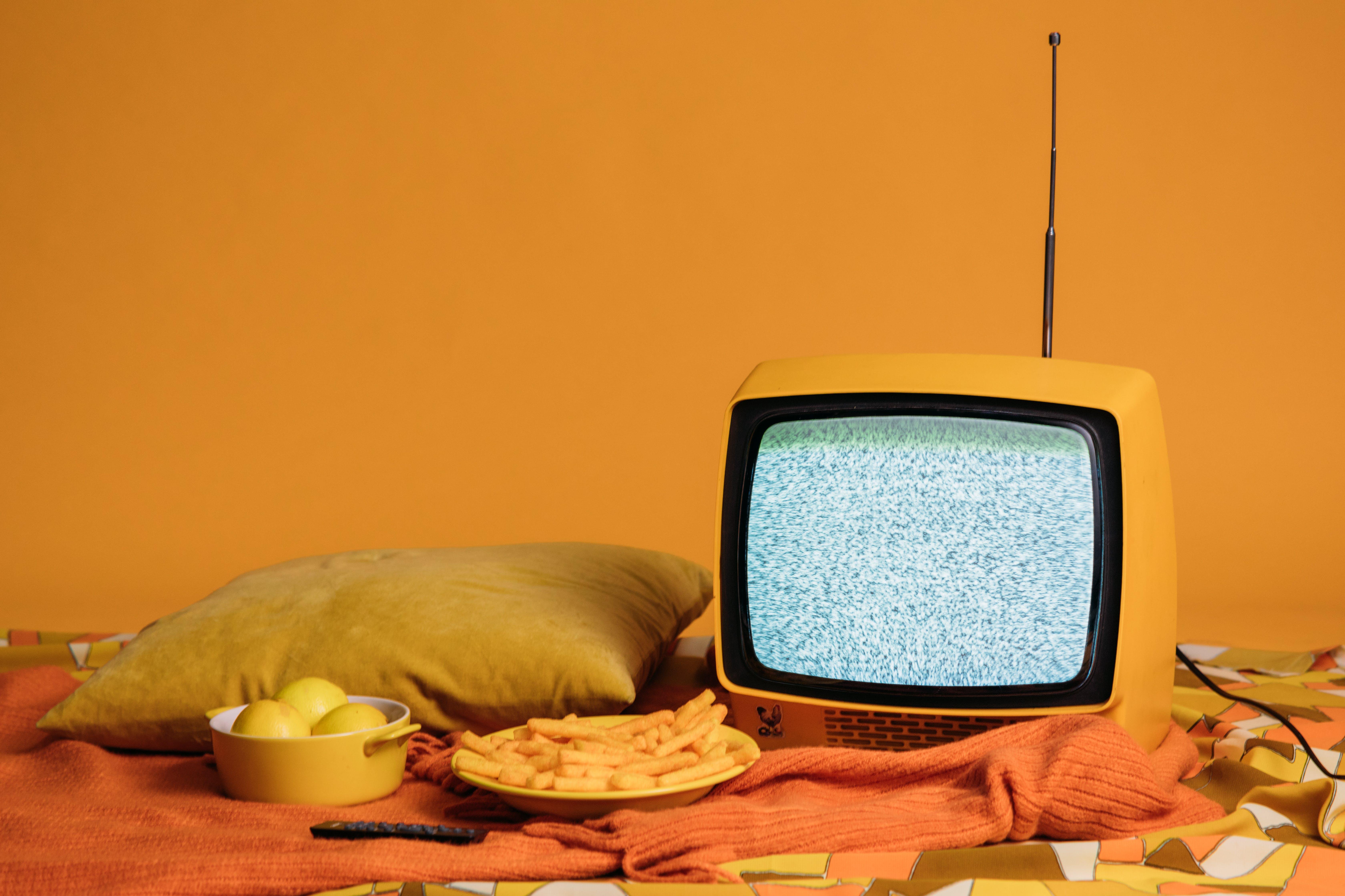 A vibrant yellow television on a bed displaying a TV advertising related content, accompanied by a bowl of fresh lemons and a plate of delicious french fries, symbolizing the blend of traditional and digital media consumption.